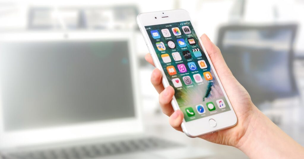 Close-up view of a smartphone displaying apps, held by a hand, with a blurred laptop in the background.